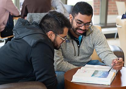 A student tutoring another while looking at an open book.